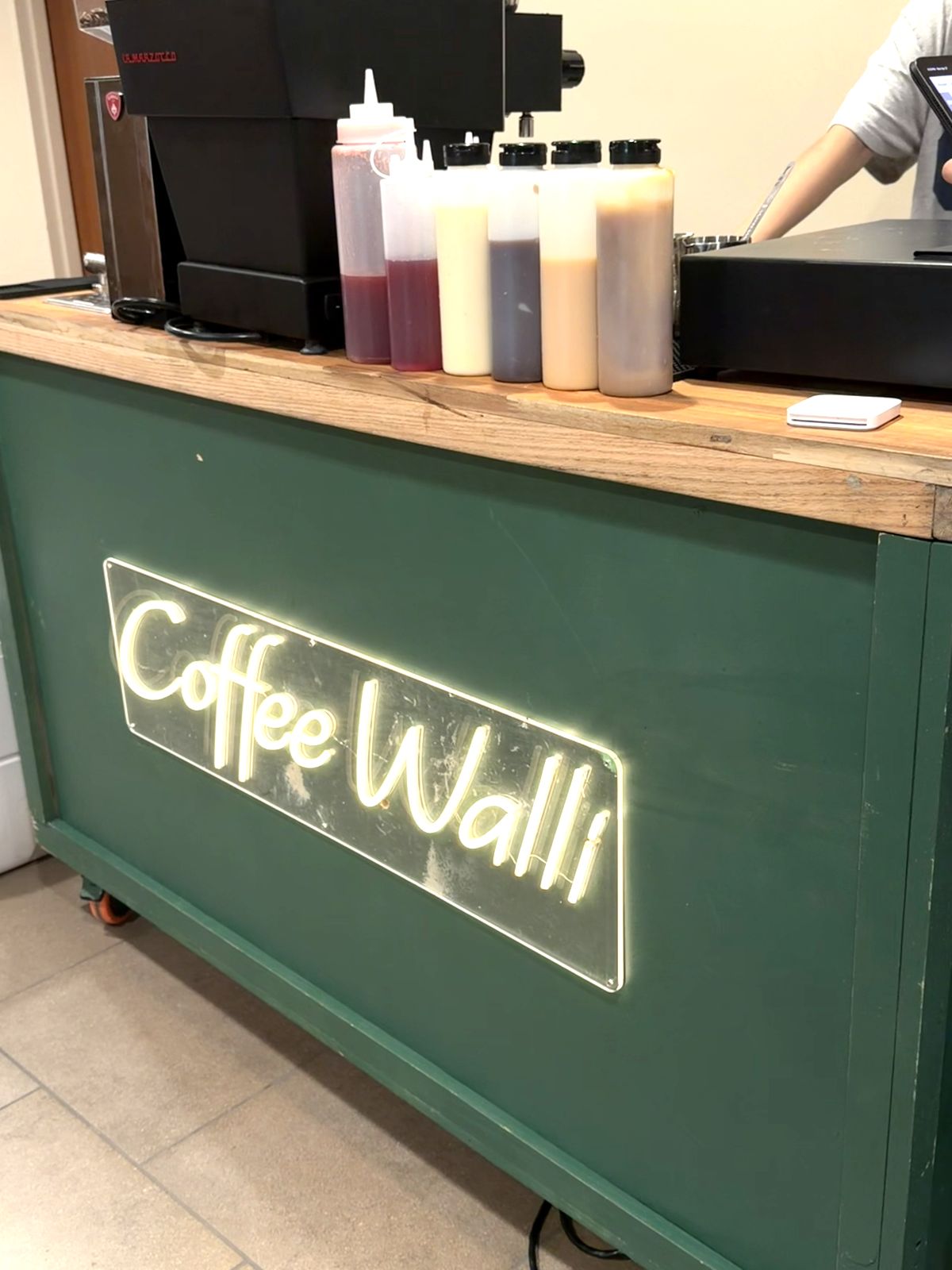 The green Coffee Walli stand with espresso machine, house-made sauce bottles, and neon sign