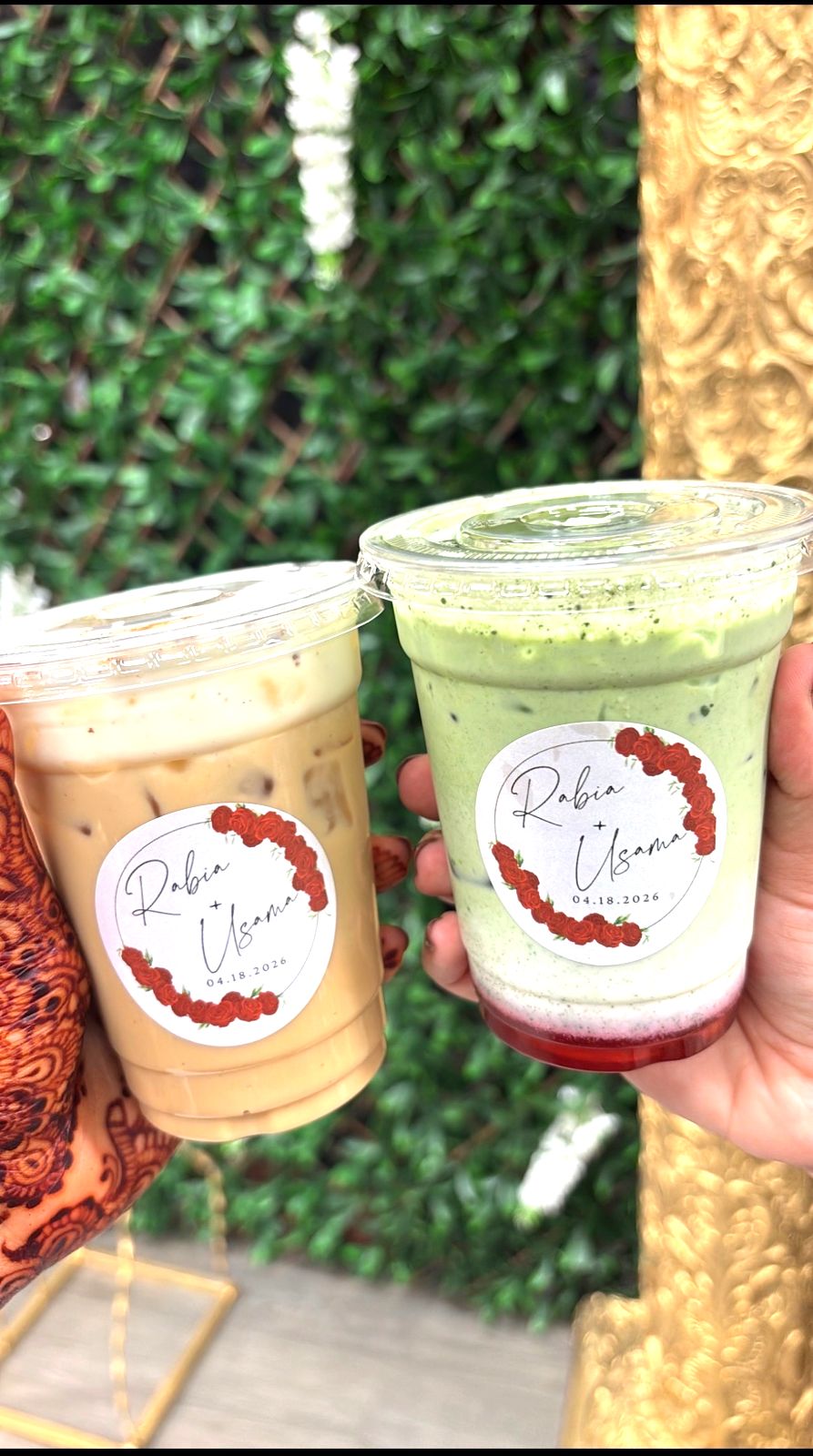 Two custom wedding drinks, an iced latte and a strawberry matcha with personalized labels, held by henna-decorated hands
