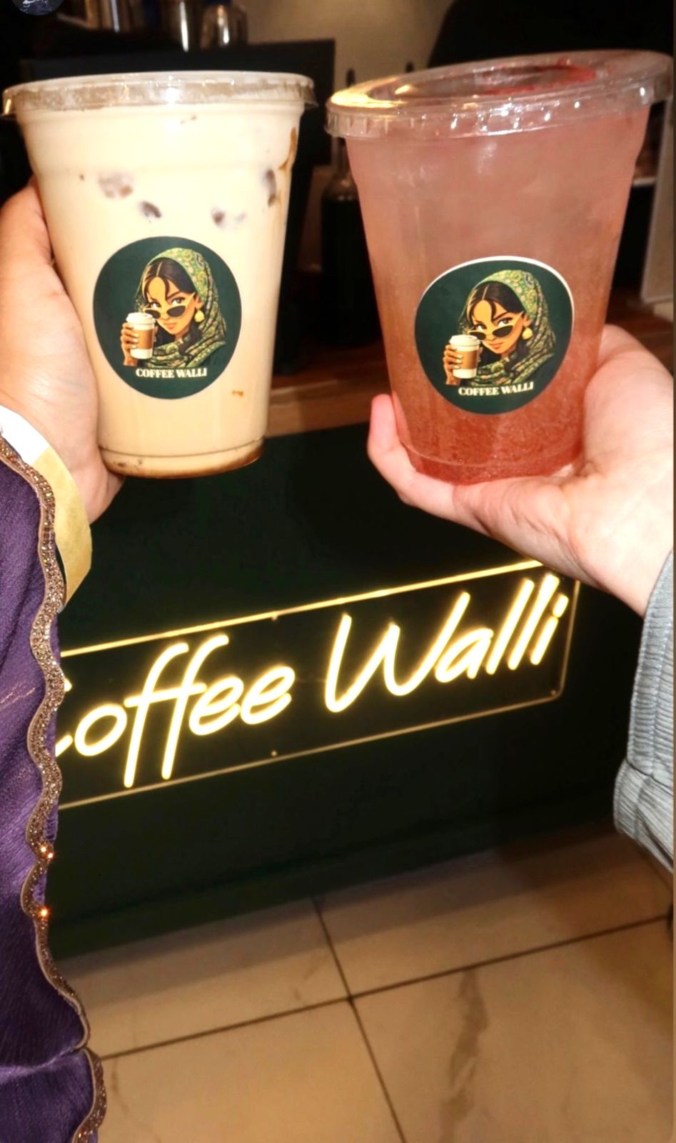 Two guests holding an iced latte and a pink sparkling refresher in front of the glowing Coffee Walli neon sign