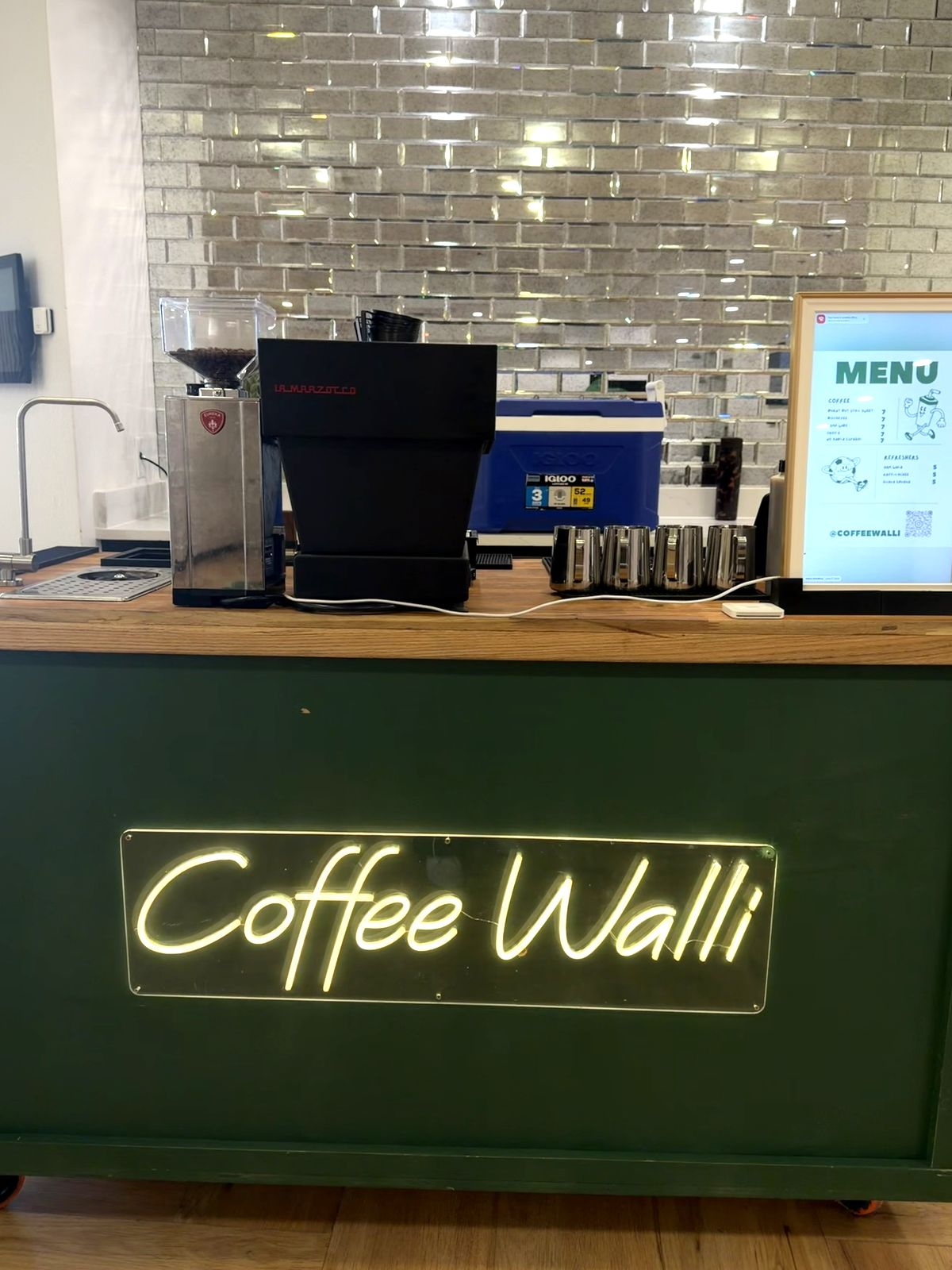 The full Coffee Walli setup with espresso machine, grinder, digital menu screen, and neon sign on the green counter